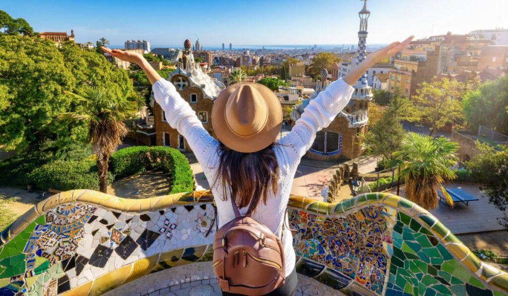 O femeie , turista, cu spatele, cu o palarie pe cap si ghiozdan in spate, cu mainile ridicate, larg deschise, in Park Güell, Barcelona, Spaina, intr-o zi insorita