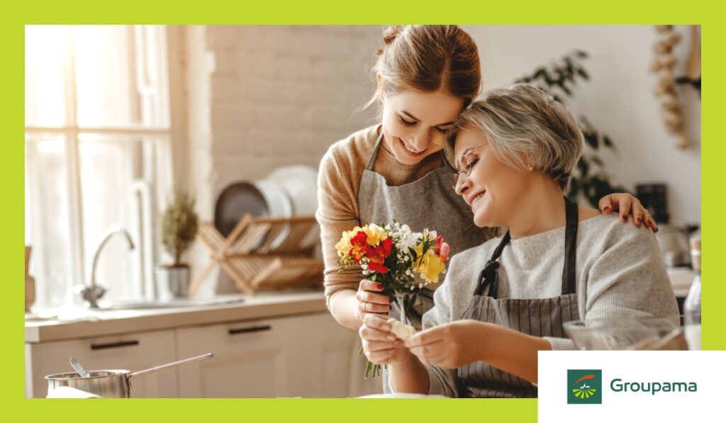 O mama impreuna cu fiica. Mama, cu parul carunt sta jos pe scaun, sprijinindu-si capul de umarul fiicei ei, care ii ofera un buchet de flori