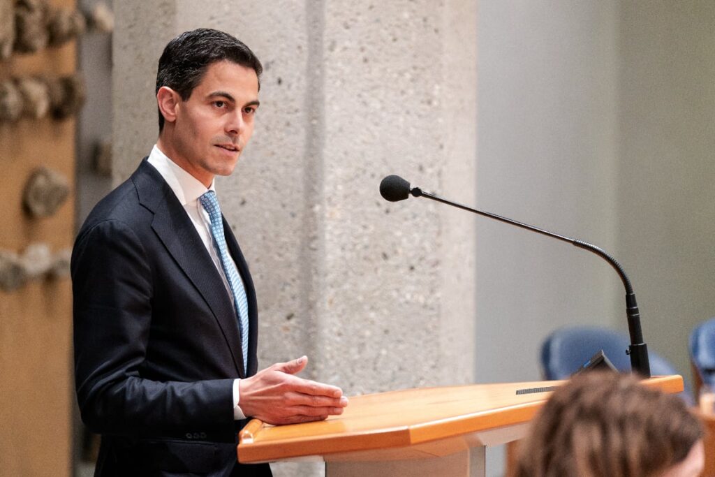 Un bărbat îmbrăcat într-un costum formal închis la culoare și o cravată albastru deschis stă la un pupitru din lemn și vorbește la un microfon. Scena sugerează un cadru instituțional sau politic, cu un fundal compus dintr-un stâlp masiv de beton și scaune cu spătar albastru, în timp ce bărbatul gesticulează cu mâinile pentru a-și sublinia discursul.