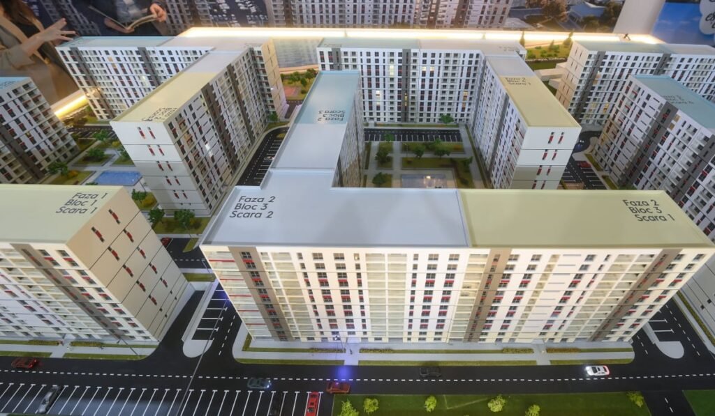 Imaginea arată o machetă arhitecturală a unui ansamblu rezidențial format din mai multe blocuri de apartamente. Blocurile sunt dispuse în jurul unor curți interioare cu spații verzi și locuri de parcare