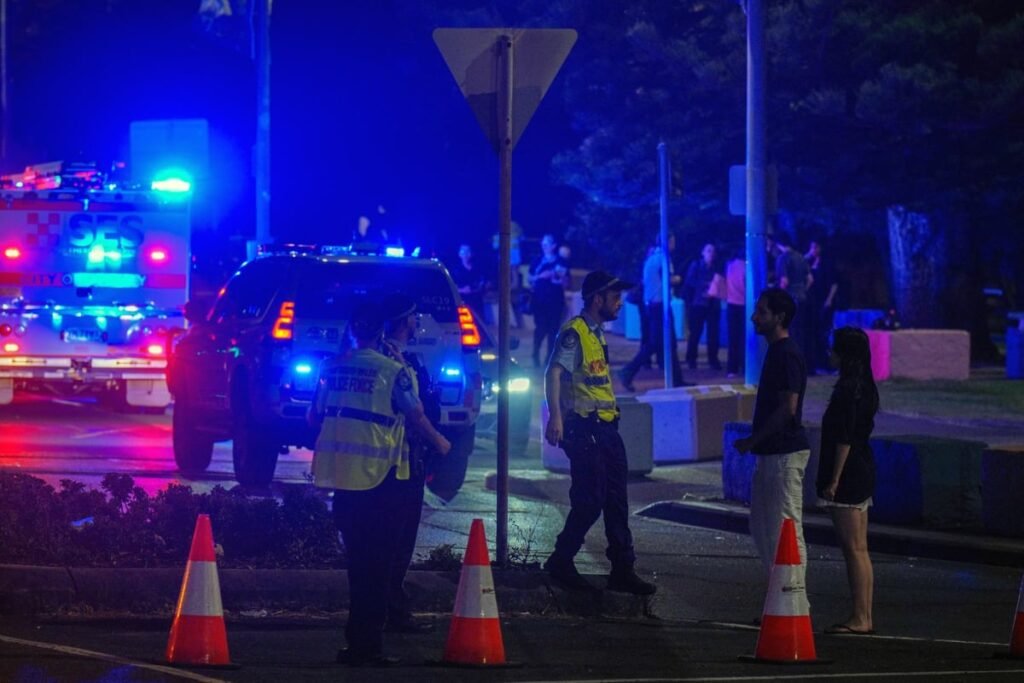 Imaginea surprinde o intervenție a forțelor de ordine pe timp de noapte, într-o zonă urbană din Australia. Polițiști și echipaje de urgență, cu girofarurile aprinse, au delimitat perimetrul cu conuri reflectorizante, în timp ce discută cu civili aflați la fața locului, sugerând o situație de securitate sau un incident grav în desfășurare.